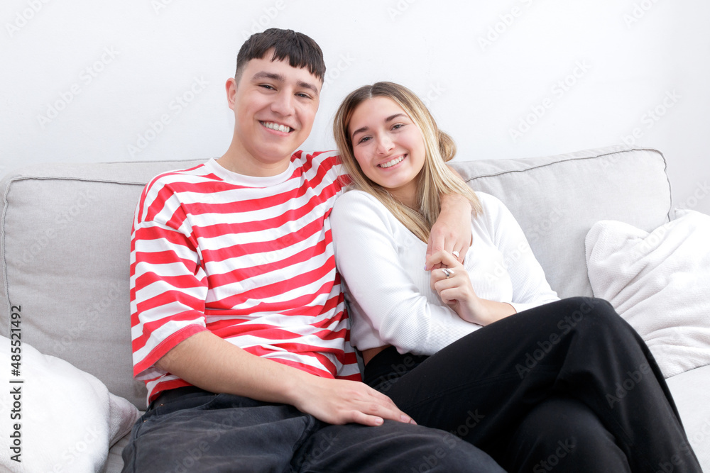 Happy embraced boy and girl sitting in the sofa smiling looking at camera. Young couple at home. Heterosexual 18-20 years old couple. Happy embraced boy and girl sitting in the sofa smiling looking at camera. Young couple at home. Heterosexual 18-20 years old couple.