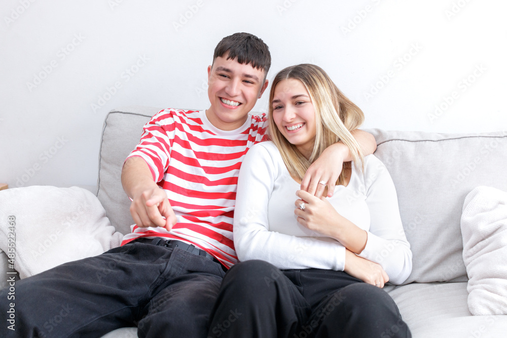 Boyfriend and girlfriend watching TV smiling. Guy pointing to the screen. Young couple at home. Heterosexual 18-20 years old couple.