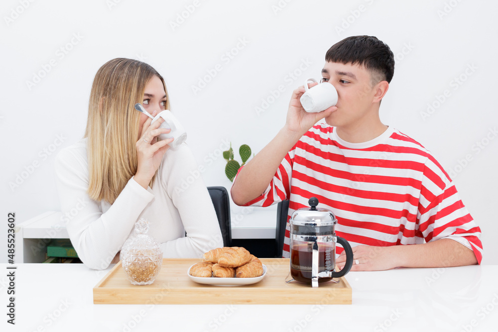 Boyfriend and girlfriend having breakfast, eating croissants and drinking coffee. Looking at each other. Young couple at home. Heterosexual 18-20 years old couple. Boyfriend and girlfriend having breakfast, eating croissants and drinking coffee. Looking at each other. Young couple at home. Heterosexual 18-20 years old couple.
