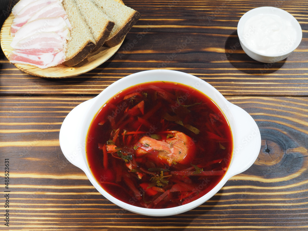Delicious hot beetroot soup in a white bowl, sour cream, rye bread with bacon on a wooden table, top view, flat layout. Traditional red borscht with meat and vegetables Delicious hot beetroot soup in a white bowl, sour cream, rye bread with bacon on a wooden table, top view, flat layout. Traditional red borscht with meat and vegetables