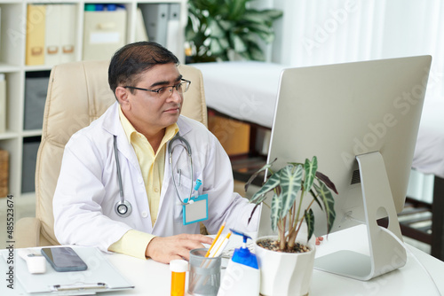 General practitioner in glasses working on laptop at his desk  he is filling medical history of patient