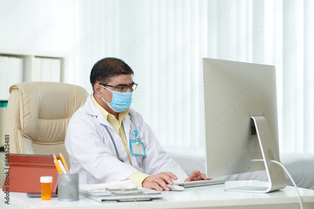 Serious chief physician in medical mask working on computer at his desk, reading documents, emails and news on coronavirus Serious chief physician in medical mask working on computer at his desk, reading documents, emails and news on coronavirus