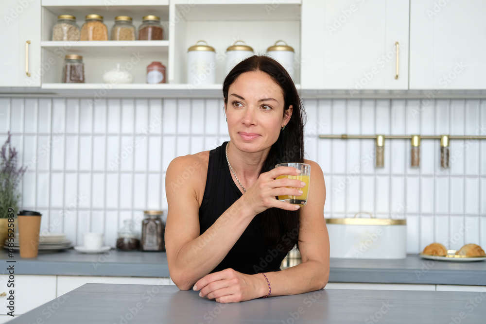 Yoga classes at home. A slender woman after a workout at home in a tracksuit stands in the kitchen and drinks juice from a glass and looks to the left Yoga classes at home. A slender woman after a workout at home in a tracksuit stands in the kitchen and drinks juice from a glass and looks to the left