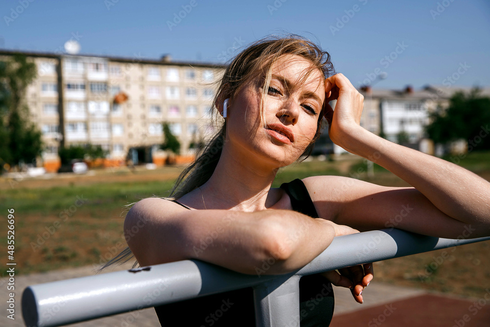 Portrait of a girl in a nasty uniform and wireless headphones looks at the camera leaning on the bars in the harsh sunlight.