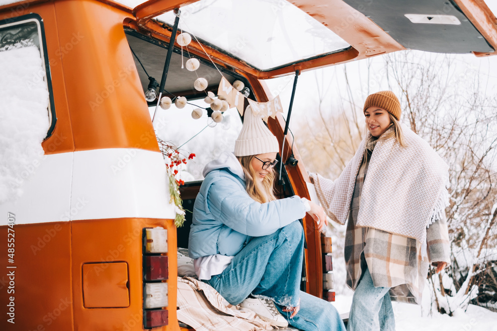 Portrait of two cheerful women friends sitting in a van in winter camp, enjoying holiday.