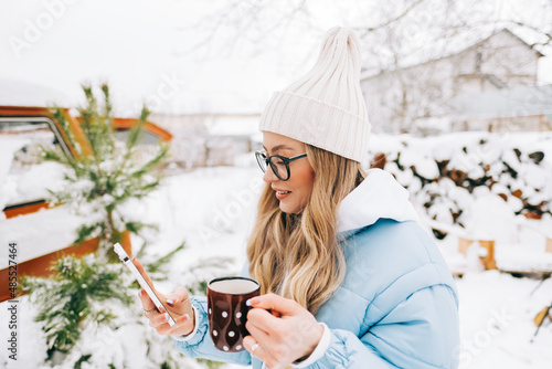 Young caucasian woman drinking tea and using mobile phone on the backyard in winter time.