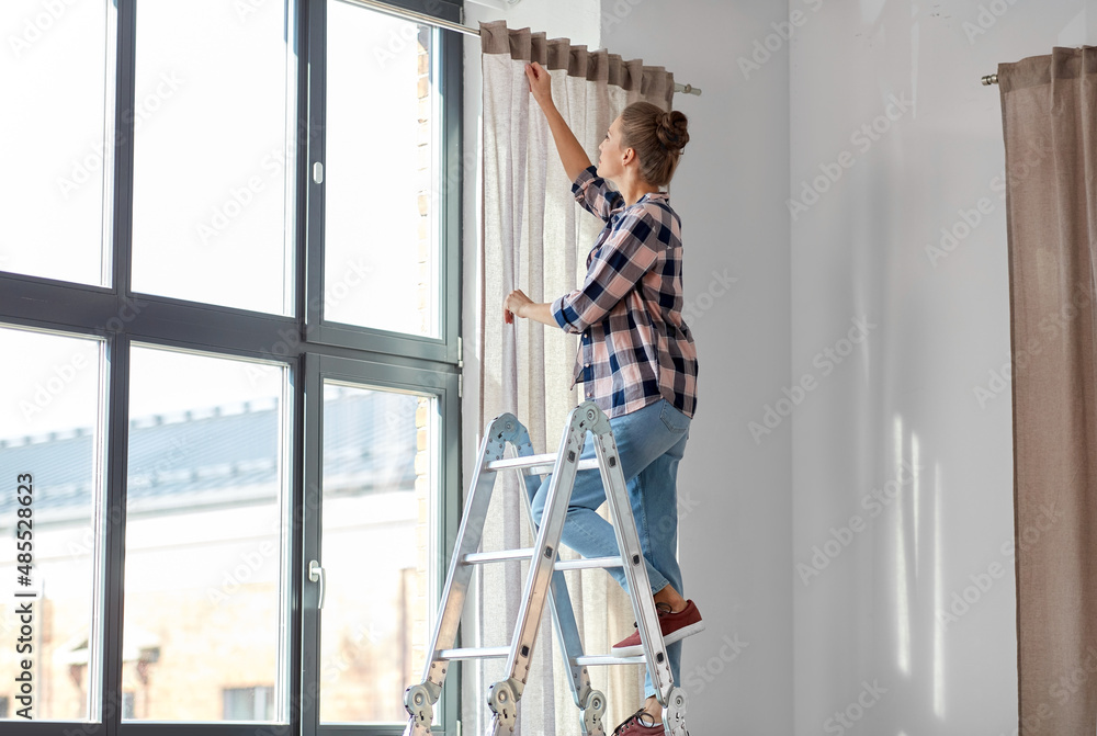 home improvement, decoration and renovation concept - happy smiling woman on ladder hanging curtains home improvement, decoration and renovation concept - happy smiling woman on ladder hanging curtains