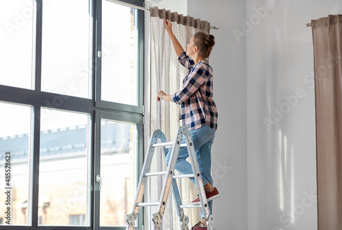 home improvement, decoration and renovation concept - happy smiling woman on ladder hanging curtains