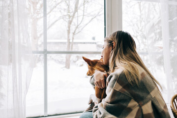 Young caucasian happy woman sitting near window with her dog in winter holidays, looking outside.