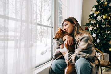 Young caucasian happy woman sitting near window with her dog in winter holidays, looking outside.