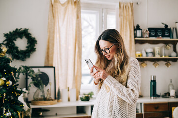 Young caucasian woman using mobile phone, standing in the kitchen with Christmas decoration at home.