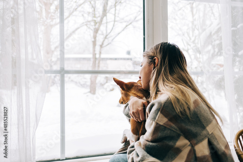 Young caucasian happy woman sitting near window with her dog in winter holidays, looking outside.
