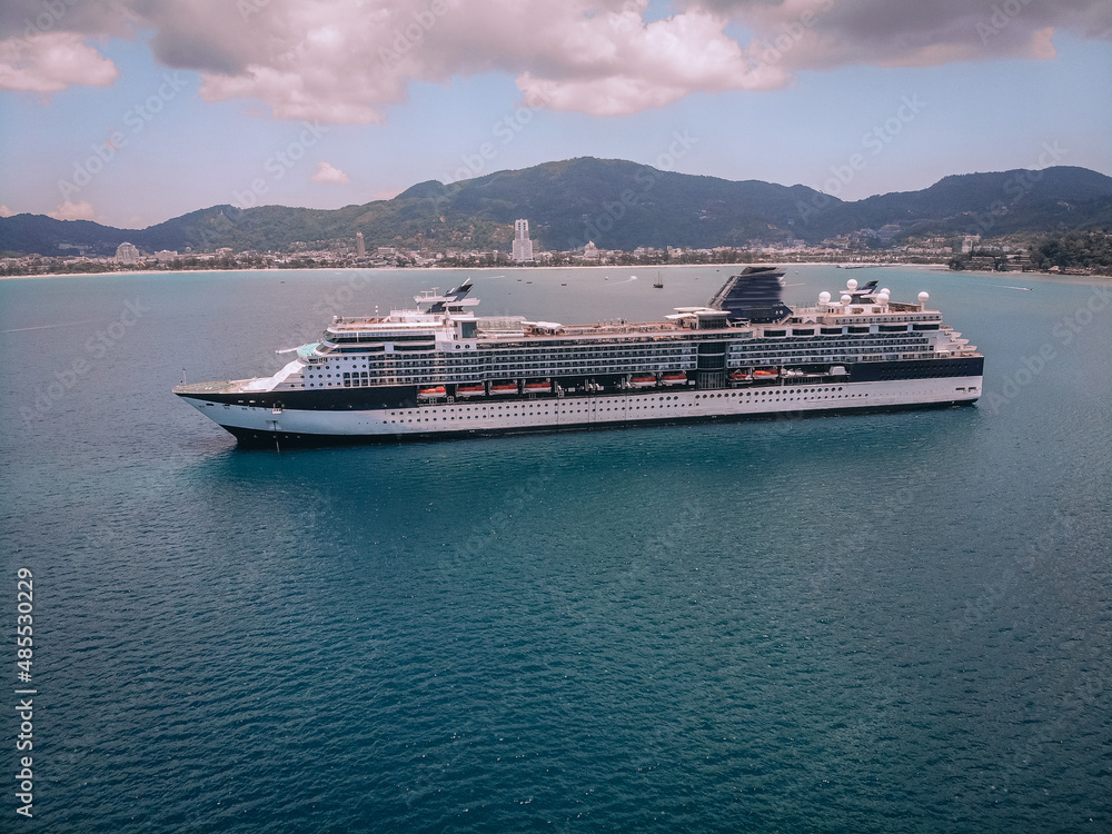 Beautiful cruise liner in the blue sea, treed slopes and coastal town on the background; luxury recreation concept.