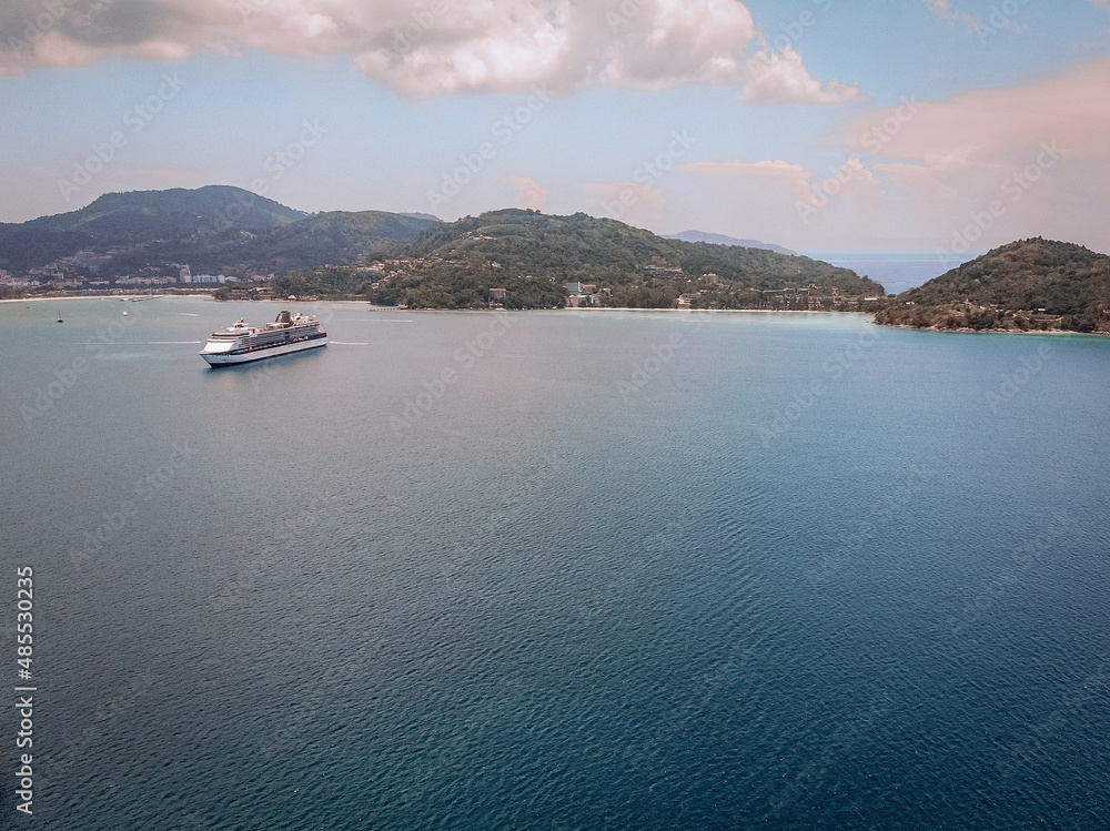Black and white liner crosses a beautiful blue bay, coastal Thai town on the background; drone shot, side view.