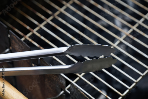 Stainless steel grill tongs on a dirty grill rust. Wooden handle blurred in the foreground.