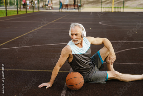 Senior man listening music while sitting with ball on playground