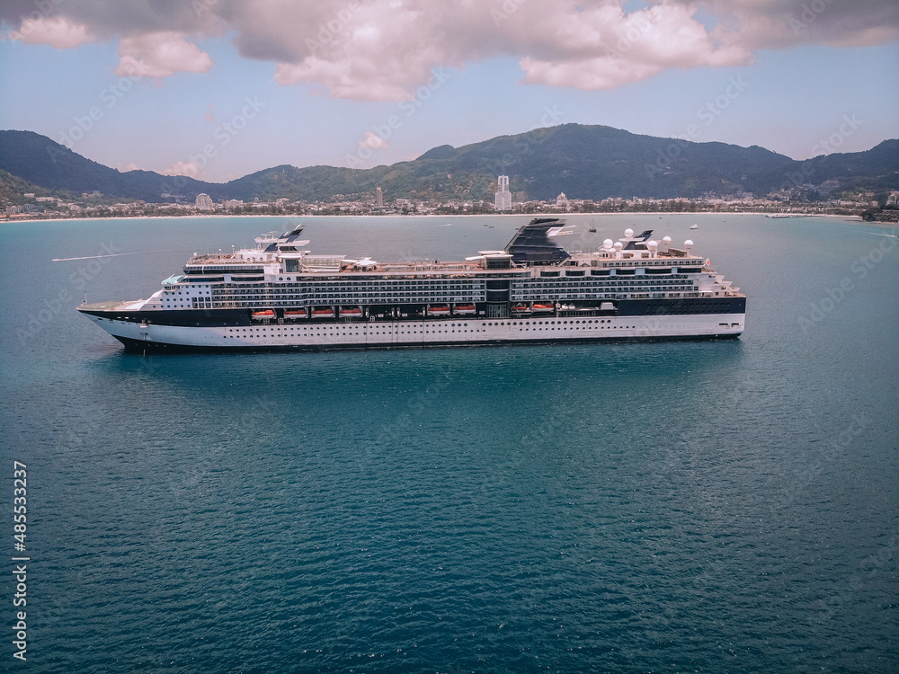 Beautiful cruise liner in the blue sea, treed slopes and coastal town on the background; luxury recreation concept.