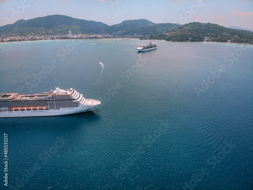 Huge white cruise liner is crossing the bay, coastal city and treed slopes on the background. Scenic harbor and blue water; drone view.