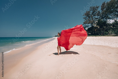 Gorgeous girl in the sunny exotic beach by the ocean. The young woman wears amazing red dress aflutter in the light breeze  backside view  vogue concept.