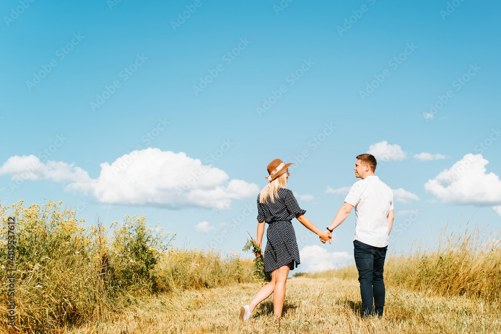 Back view of happy couple holding hands in meadow and blue sky outdoors. Cute young caucasian family spending time together in nature on sunny summer day. Romantic date, valentine's day, love story