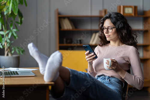 Woman freelance writer rest or procrastinate at workplace. Businesswoman in eyewear distracted from work on laptop scrolling social media on smartphone. Freelancer holding mobile phone and browsing photo