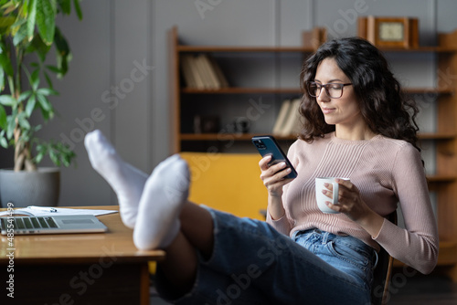 Woman freelance writer rest or procrastinate at workplace. Businesswoman in eyewear distracted from work on laptop scrolling social media on smartphone. Freelancer holding mobile phone and browsing