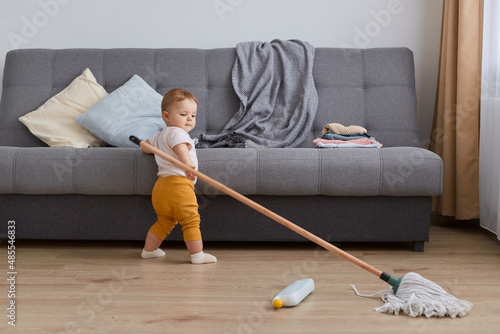 Canvas Print Indoor shot of infant toddler baby wearing yellow pants standing near gray sofa with mop, little charming girl helping mother doing household chores, try to wash floor
