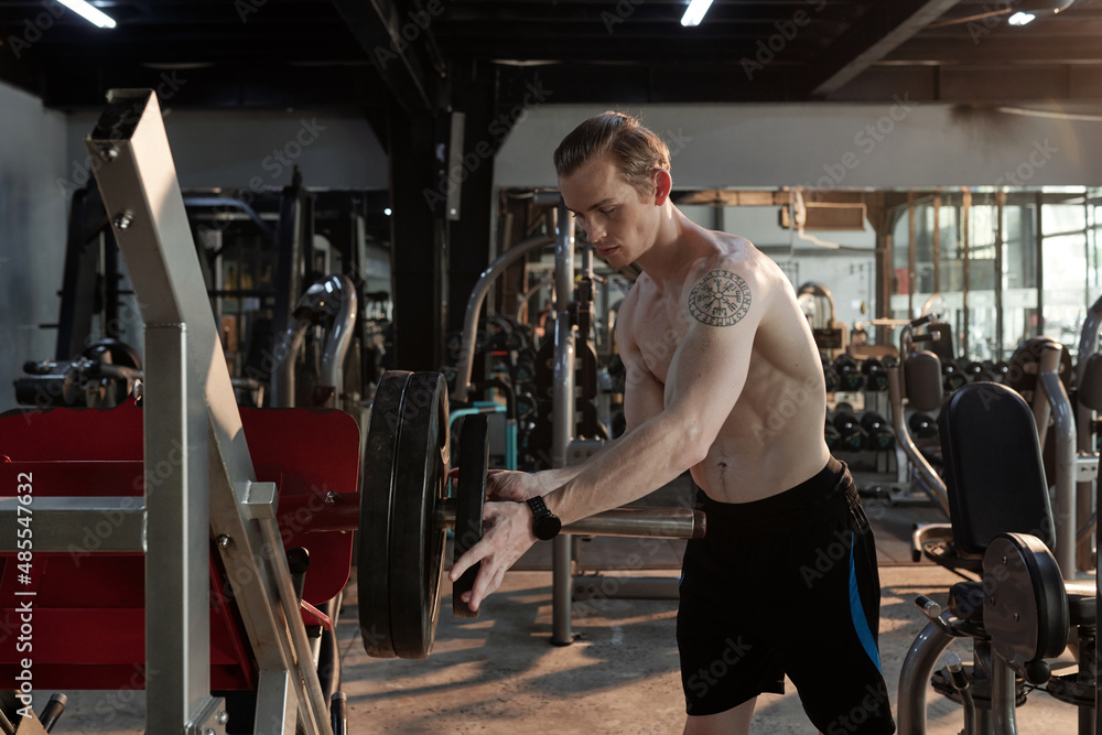 Shirtless fit young man putting weight plate on barbell in gym and getting ready for exercise Shirtless fit young man putting weight plate on barbell in gym and getting ready for exercise