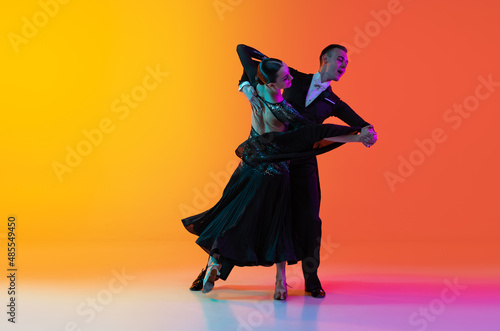 Dynamic portrait of young beautiful man and woman dancing ballroom dance isolated over gradient orange pink background in neon light. Beauty, art, sport concept