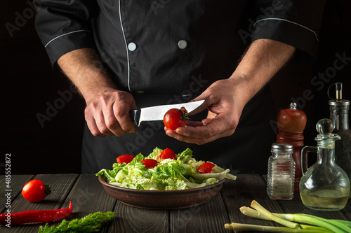 The chef prepares a vegetable salad in the restaurant kitchen. Cutting a fresh tomato for a vitamin salad with a knife