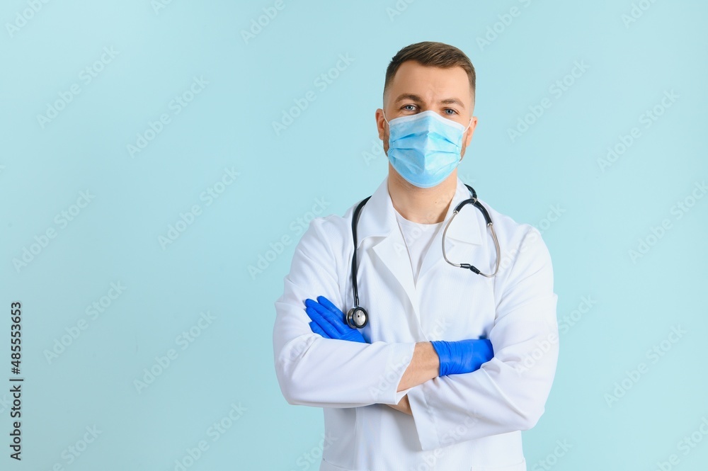 Young male doctor wearing medical mask, and stethoscope round his neck, isolated on blue background