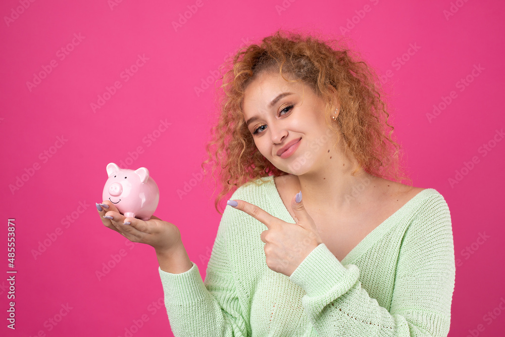 A cute young girl with curly red hair holds a piggy bank, a pink piglet in her hands. The concept of wealth and safety of money. A cute young girl with curly red hair holds a piggy bank, a pink piglet in her hands. The concept of wealth and safety of money.