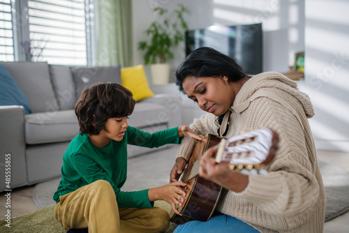 Little multiracial boy learning to play the guitar with his mother at home.