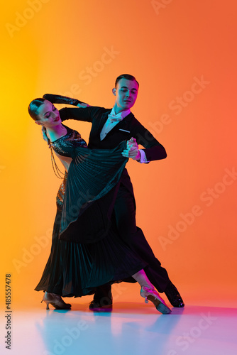 Full-length portrait of young beautiful man and woman dancing ballroom dance isolated over gradient orange pink background in neon light. Beauty, art, sport concept