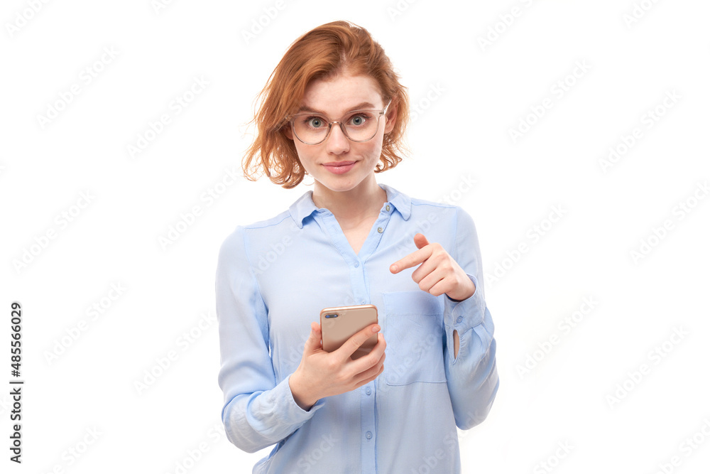 Shocked joyful redhead girl points finger at smartphone blank screen in glasses and business shirt isolated on white studio background Shocked joyful redhead girl points finger at smartphone blank screen in glasses and business shirt isolated on white studio background