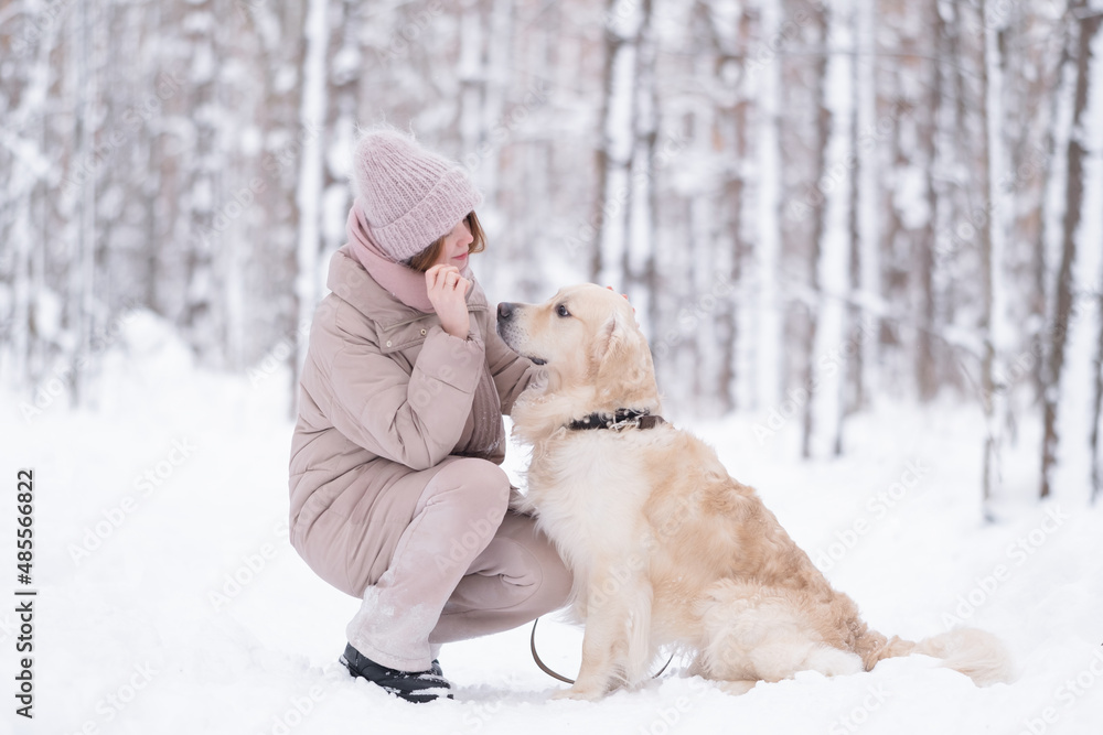 A young redheaded woman plays with her dog in the winter woods. A beautiful girl walks through a picturesque snow-covered park with a golden retriever. The concept of friendship with a pet.