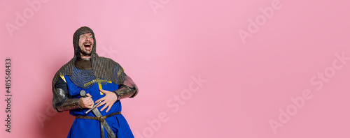 Portrait of laughing man, medieval warrior or knight in armor with sword isolated over pink studio background. Flyer
