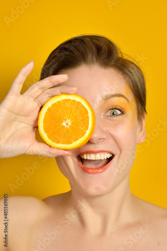 A young beautiful woman with short hair, bright makeup on a yellow background, holding a juicy orange in her hands and smiling. Funny funny attractive model with a slice of orange in her hand