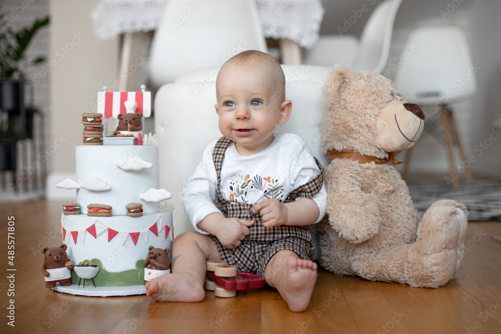 tiny adorable child sitting on floor near the birthday cake and teddy bear. Looking with sad eyes. Close-up photo tiny adorable child sitting on floor near the birthday cake and teddy bear. Looking with sad eyes. Close-up photo