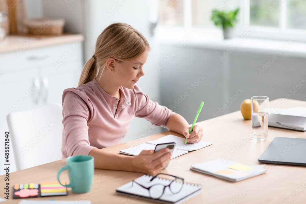 Serious busy pretty caucasian teenage girl blonde makes notes with smartphone at table in minimalist kitchen Serious busy pretty caucasian teenage girl blonde makes notes with smartphone at table in minimalist kitchen