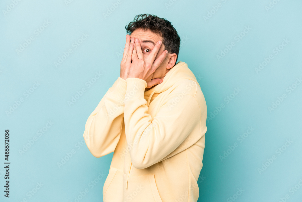 Young caucasian man isolated on blue background blink through fingers frightened and nervous. Young caucasian man isolated on blue background blink through fingers frightened and nervous.