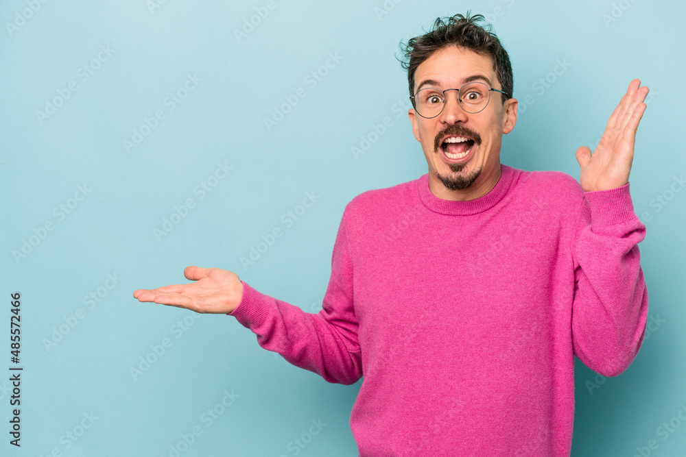 Young caucasian man isolated on blue background holds copy space on a palm, keep hand over cheek. Amazed and delighted. Young caucasian man isolated on blue background holds copy space on a palm, keep hand over cheek. Amazed and delighted.