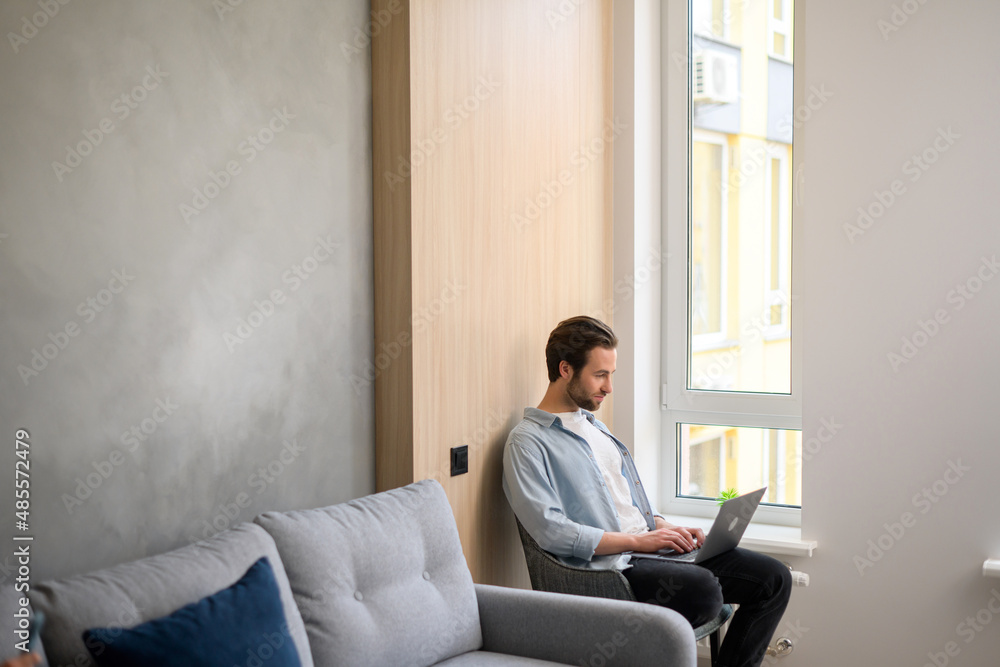 Handsome guy spends time in networks, young man sitting on the armchair in modern apartment and using a laptop, watching movies, male freelancer working remotely from home, typing Handsome guy spends time in networks, young man sitting on the armchair in modern apartment and using a laptop, watching movies, male freelancer working remotely from home, typing