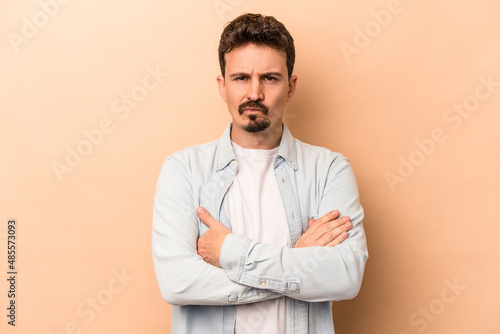 Young caucasian man isolated on beige background frowning face in displeasure, keeps arms folded. © Asier