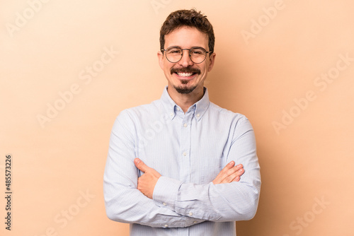 Young caucasian man isolated on beige background who feels confident, crossing arms with determination.