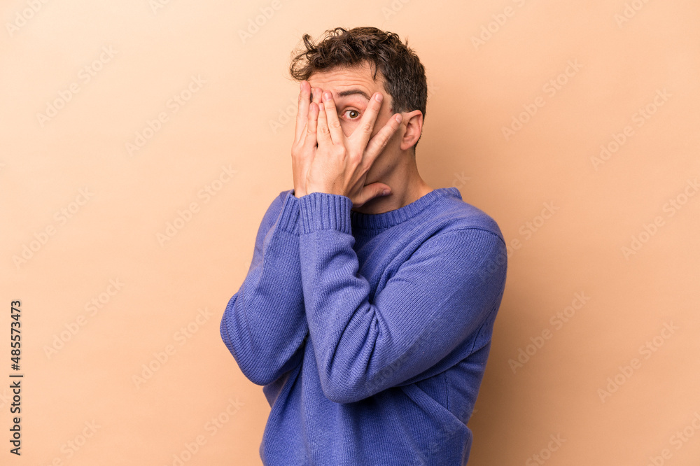 Young caucasian man isolated on beige background blink through fingers frightened and nervous. Young caucasian man isolated on beige background blink through fingers frightened and nervous.