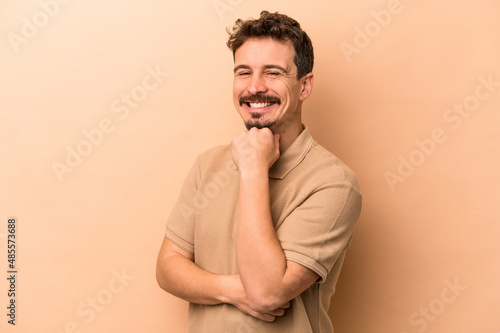 Young caucasian man isolated on beige background smiling happy and confident, touching chin with hand. © Asier