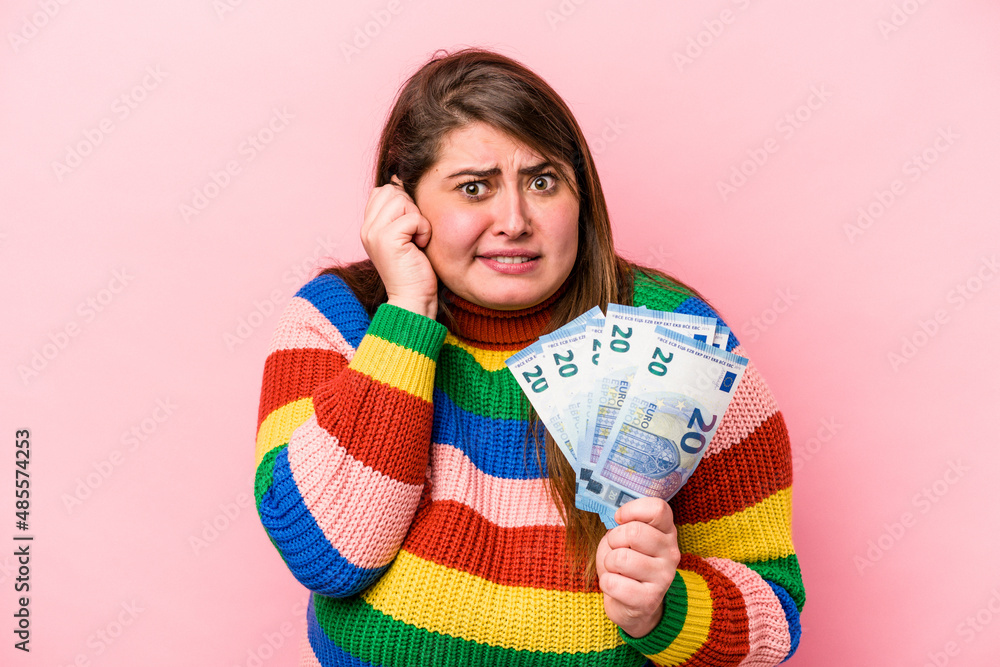 Young caucasian overweight woman holding banknotes isolated on pink background covering ears with hands. Young caucasian overweight woman holding banknotes isolated on pink background covering ears with hands.