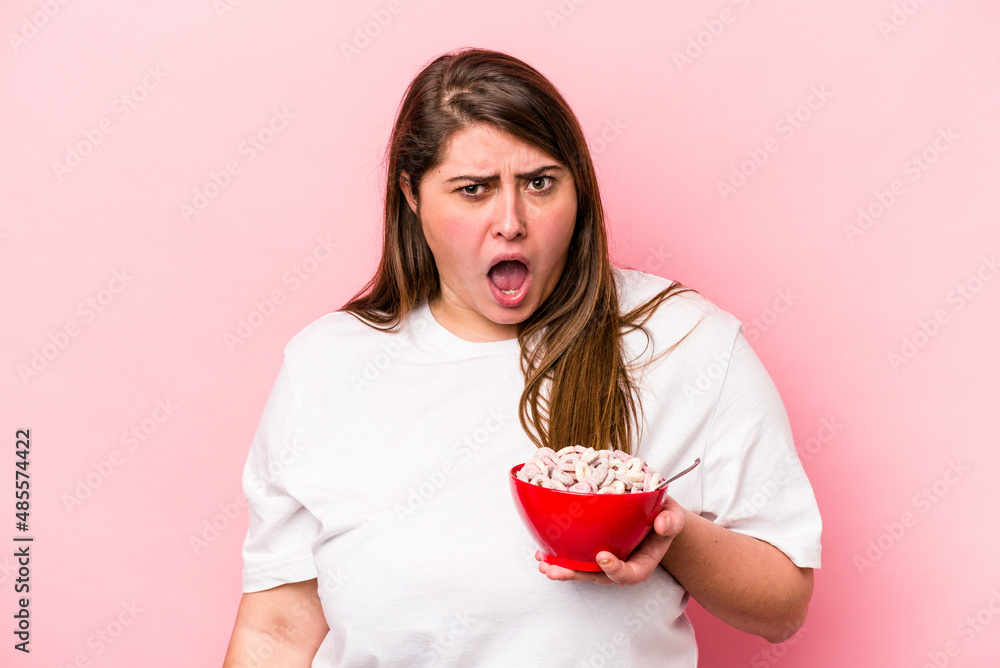 Young caucasian overweight woman holding a bowl of cereals isolated on pink background screaming very angry and aggressive. Young caucasian overweight woman holding a bowl of cereals isolated on pink background screaming very angry and aggressive.
