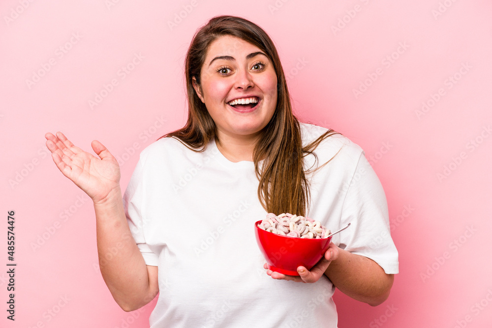 Young caucasian overweight woman holding a bowl of cereals isolated on pink background receiving a pleasant surprise, excited and raising hands. Young caucasian overweight woman holding a bowl of cereals isolated on pink background receiving a pleasant surprise, excited and raising hands.